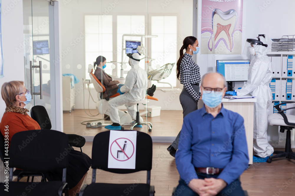 Obraz premium Dentist nurse dressed in ppe suit with face shiled discussing with patient in stomatology waiting room. People keeping social distancing as prevention during coronavirus outbreak.