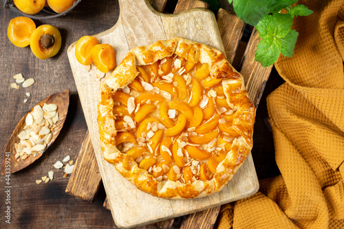 French Galette or a pie with apricots and almonds on on a wooden serving board on a brown culinary background top view. Summer homemade sweet pastries photo