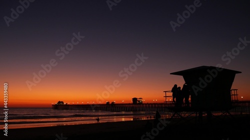 Young teen girls silhouettes near lifeguard tower, friends on pacific ocean beach, sunset dusk in Oceanside, California USA. Unrecognizable teenagers, people and twilight gradient purple violet sky. © Dogora Sun