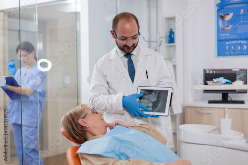 Stomatolog in uniform during consultation of senior woman explaining diagnostic. Medical teeth care taker holding patient radiography on tablet pc near patient standing up.