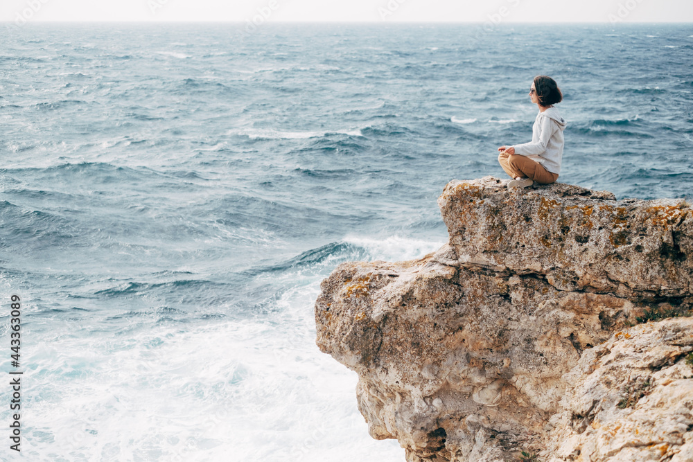 Young woman sits on the edge of a rocky shore in a lotus position.