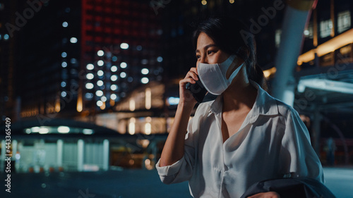 Serious dissatisfied upset young Asia businesswoman wear medical face mask talk via phone while walk alone outdoor in urban city night. Business on go, Social distancing to prevent spread of COVID-19. © tirachard