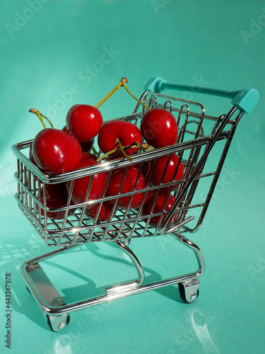 mini metal supermarket cart filled with red juicy cherries on a green background side view