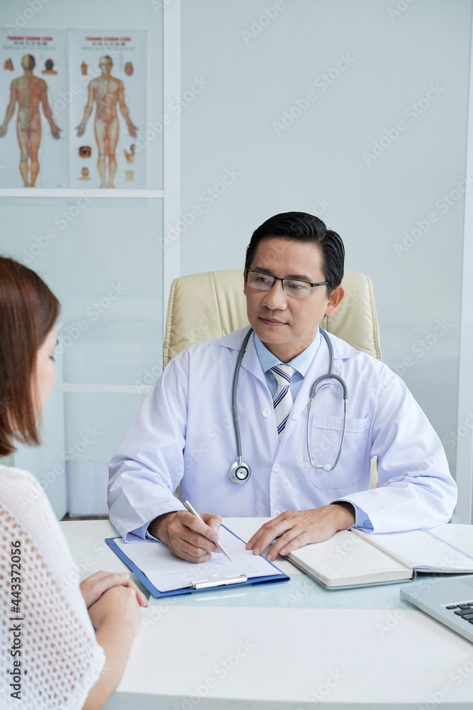 Fototapeta premium Serious doctor listening to young female patient and taking notes in document