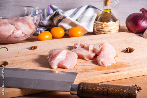 Sliced pieces of raw chicken breast, spices and ingredients for cooking oriental chicken dish on a cutting board with a Chinese knife.
