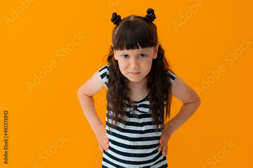 angry child is a cute 5-6-year-old girl in a striped dress on an isolated yellow background. Capricious transitional age self-affirmation.