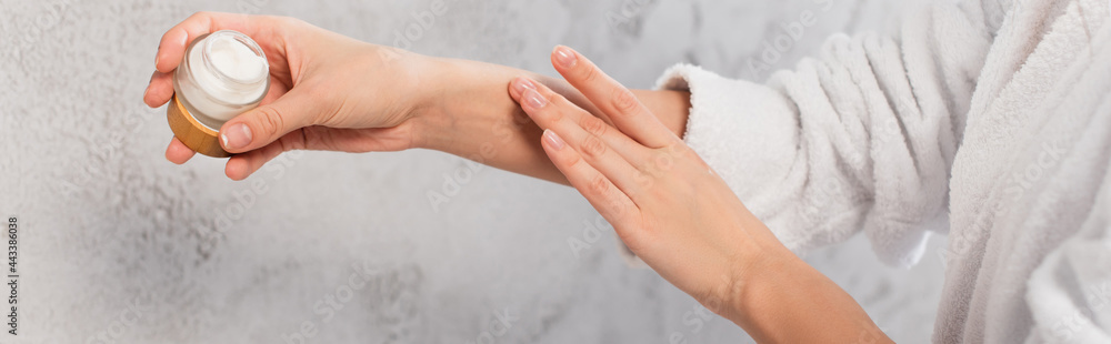 Naklejka premium cropped view of young woman in bathrobe holding container and applying cosmetic cream, banner