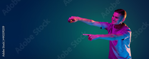 Young Caucasian man in casual clothes isolated over gradient blue green studio background in pink neon light with copyspace for ad. Concept of human emotions