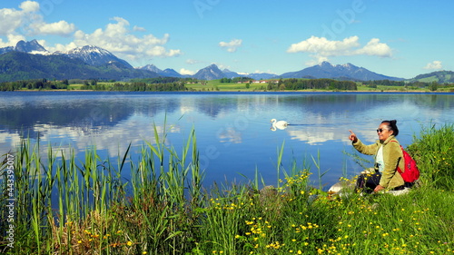 junge Frau genießt die stille Natur am Hopfensee im Allgäu am grünen Ufer sitzend bei blauem Himmel 