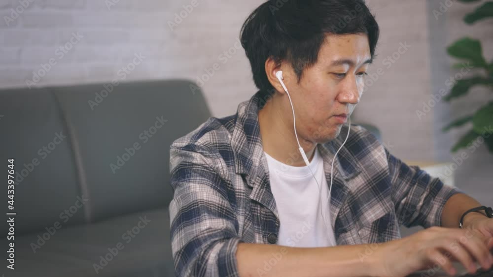 Young Asian male entrepreneur in casual clothing and earphone working on computer laptop at late night from home Young Asian male entrepreneur in casual clothing and earphone working on computer laptop at late night from home