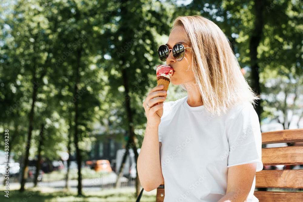 Attractive young woman with glasses resting on a park bench and eating ice cream in a waffle cone. Beautiful blonde girl chill summer time, outdoors. Summer lifestyle