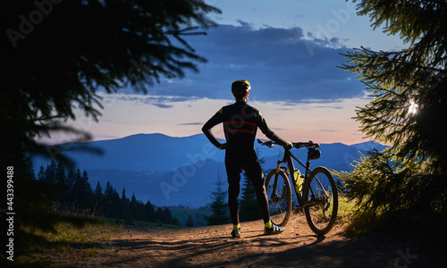 Back view on male cyclist standing holding his bike, watching beautiful landscape, mountains peaks and sunset, surrounded with spruces in the evening. Active lifestyle concept