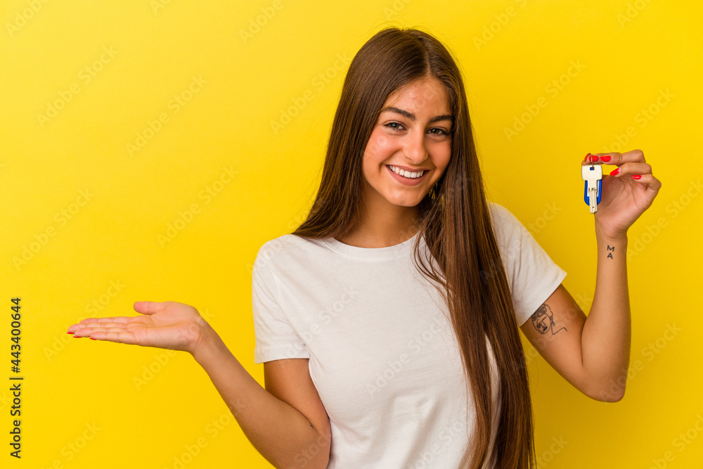 Young caucasian woman holding a home keys isolated on yellow background showing a copy space on a palm and holding another hand on waist. Young caucasian woman holding a home keys isolated on yellow background showing a copy space on a palm and holding another hand on waist.