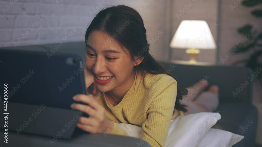 Young Asian woman relaxing at home, lying on couch on stomach with pillows using digital tablet and enjoying movies at home during late night Young Asian woman relaxing at home, lying on couch on stomach with pillows using digital tablet and enjoying movies at home during late night