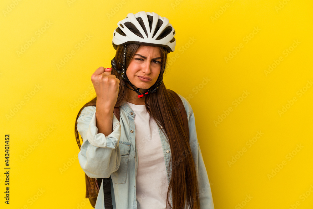 Young student caucasian woman wearing a bike helmet isolated on yellow background showing fist to camera, aggressive facial expression. Young student caucasian woman wearing a bike helmet isolated on yellow background showing fist to camera, aggressive facial expression.