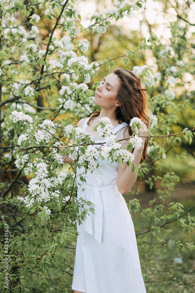 A close-up portrait of a happy pretty gentle smiling young woman with a hairstyle in a white cotton dress having fun walking alone enjoying the smell of blooming flowers in a summer green outdoor park A close-up portrait of a happy pretty gentle smiling young woman with a hairstyle in a white cotton dress having fun walking alone enjoying the smell of blooming flowers in a summer green outdoor park