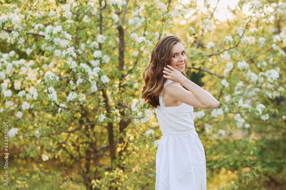 A close-up portrait of a happy pretty gentle smiling young woman with a hairstyle in a white cotton dress having fun walking alone enjoying the smell of blooming flowers in a summer green outdoor park