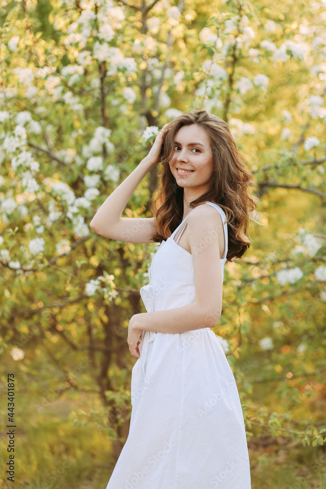 A close-up portrait of a happy pretty gentle smiling young woman with a hairstyle in a white cotton dress having fun walking alone enjoying the smell of blooming flowers in a summer green outdoor park A close-up portrait of a happy pretty gentle smiling young woman with a hairstyle in a white cotton dress having fun walking alone enjoying the smell of blooming flowers in a summer green outdoor park
