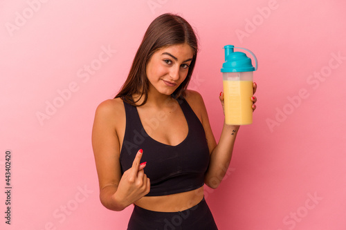 Young caucasian woman holding a protein shake isolated on yellow background pointing with finger at you as if inviting come closer.