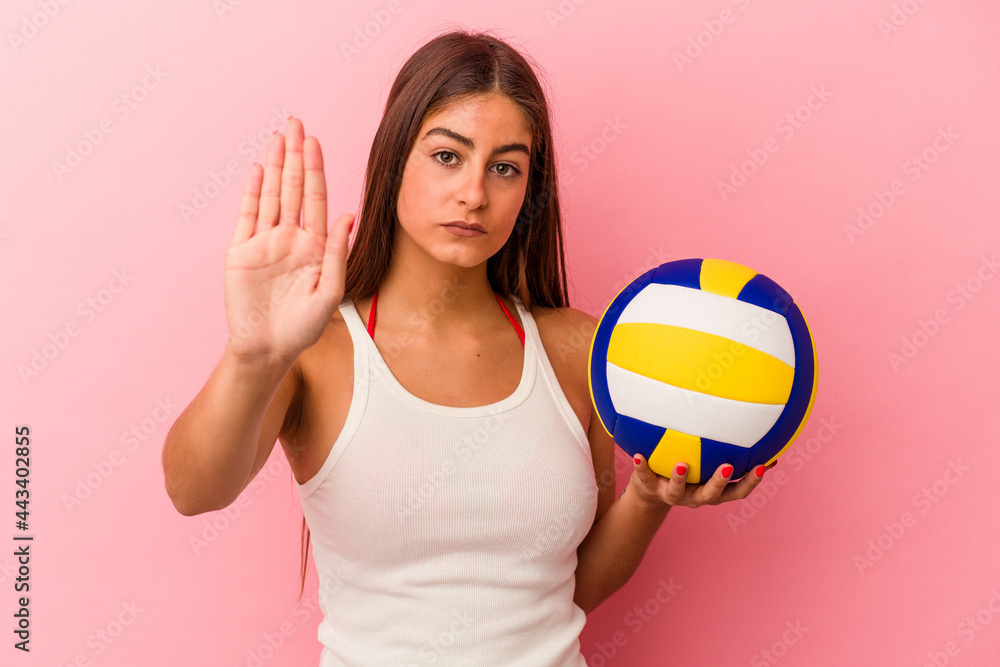 Young caucasian woman holding a volleyball ball isolated on pink background standing with outstretched hand showing stop sign, preventing you.