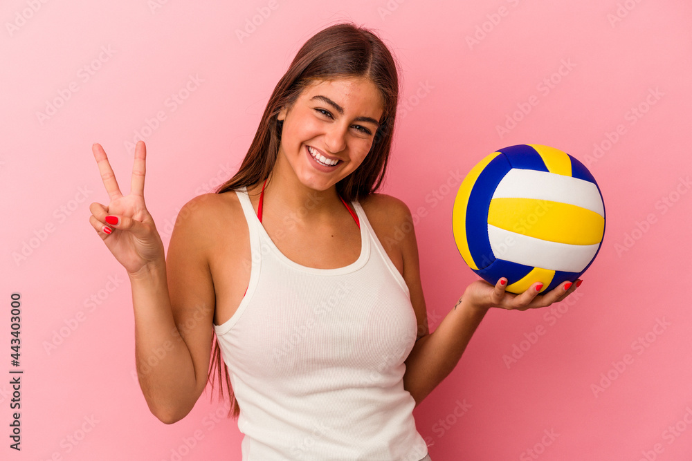 Young caucasian woman holding a volleyball ball isolated on pink background joyful and carefree showing a peace symbol with fingers.