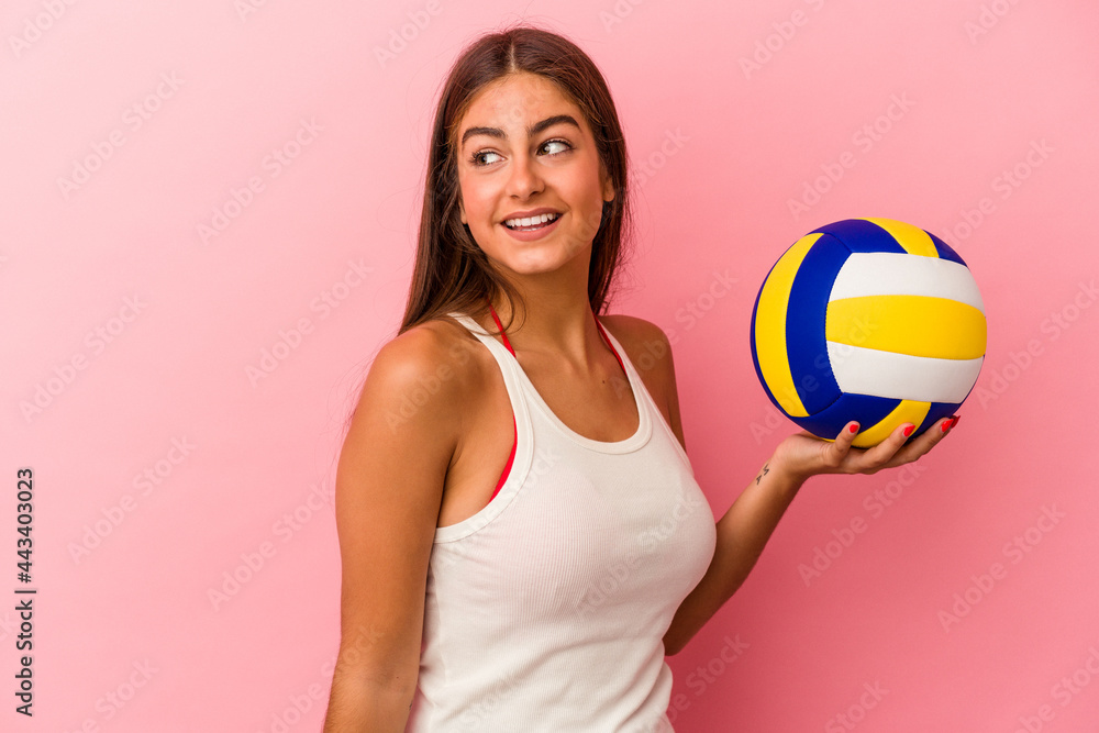 Young caucasian woman holding a volleyball ball isolated on pink background looks aside smiling, cheerful and pleasant.