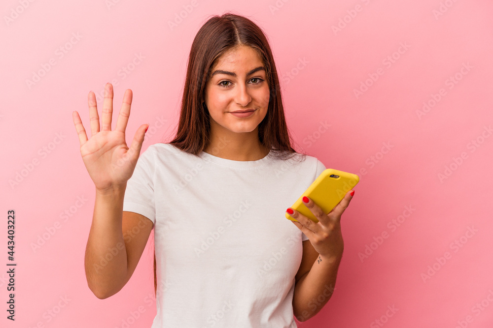Young caucasian woman holding a mobile phone isolated on pink background smiling cheerful showing number five with fingers. Young caucasian woman holding a mobile phone isolated on pink background smiling cheerful showing number five with fingers.