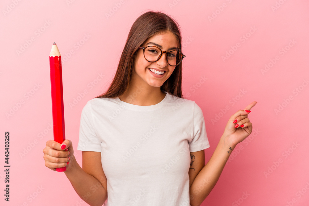 Young caucasian woman holding a big pencil isolated on pink background smiling and pointing aside, showing something at blank space. Young caucasian woman holding a big pencil isolated on pink background smiling and pointing aside, showing something at blank space.