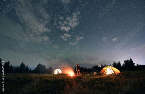 Illuminated camp tents and couple near campfire. Young woman and man travelers standing near bonfire under magical blue sky with stars. Concept of travelling  night camping and relationship.
