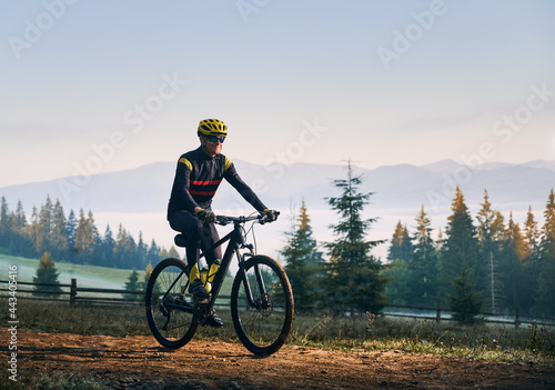 Fototapeta Naklejka Na Ścianę i Meble -  Happy cyclist in cycling suit riding bike on mountain road with coniferous trees and hills on background. Man bicyclist wearing safety helmet and glasses while enjoying bicycle ride in mountains.