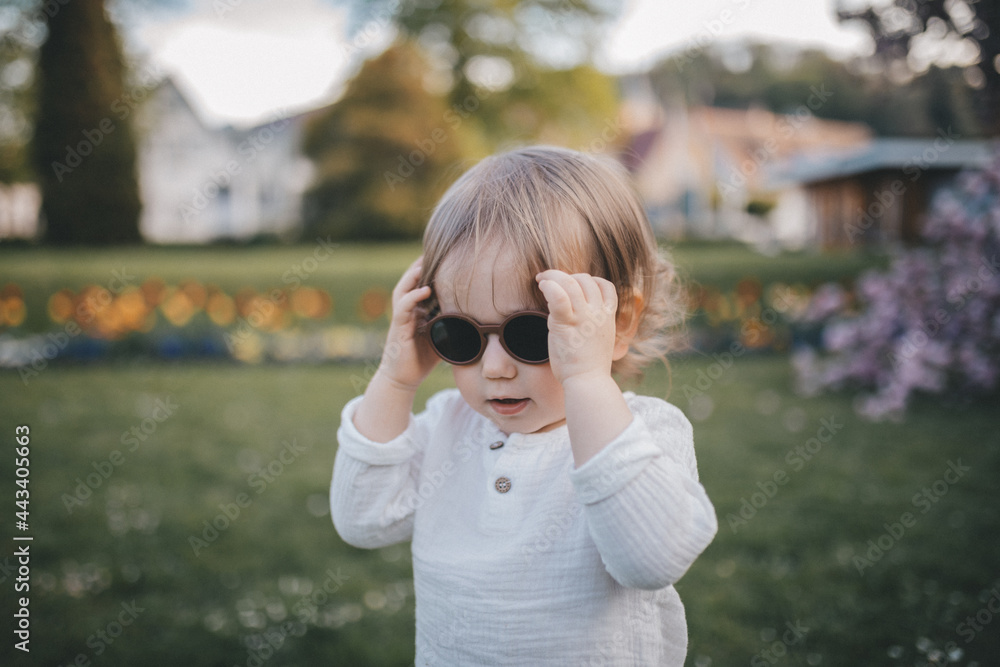 Little boy puts sunglasses on while walk in blooming garden.