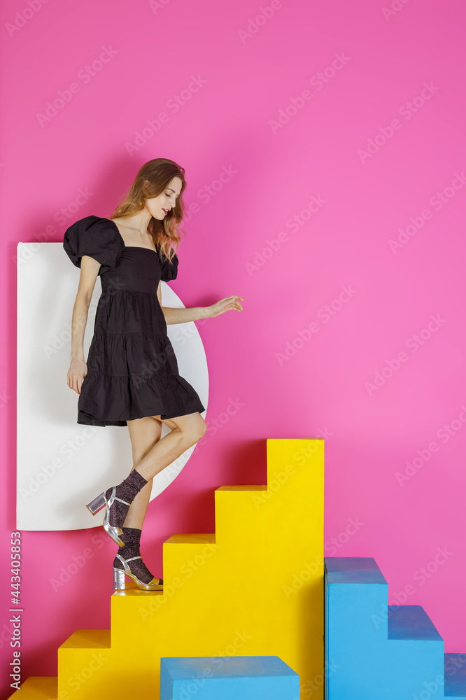 Young pleasing woman in a black dress poses in the studio against a pink wall Young pleasing woman in a black dress poses in the studio against a pink wall