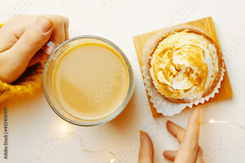 Top view of women s hands touching coffee mug and tartlet. Girl in knitted sweater is having breakfast. Romantic atmosphere. Sweet dessert and refreshing drink on white table. Still life aesthetic.