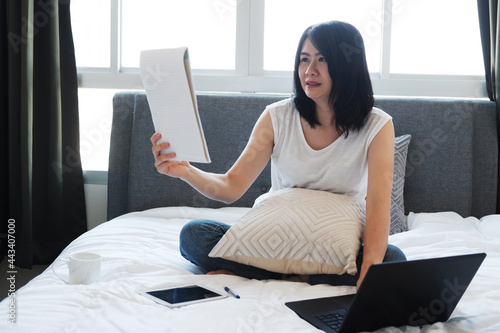 Asian young woman is sitting on white bed and reading papersheet for online working and learning with laptop in social medea in bedroom. Businesswoman work from home with Technology concept. photo