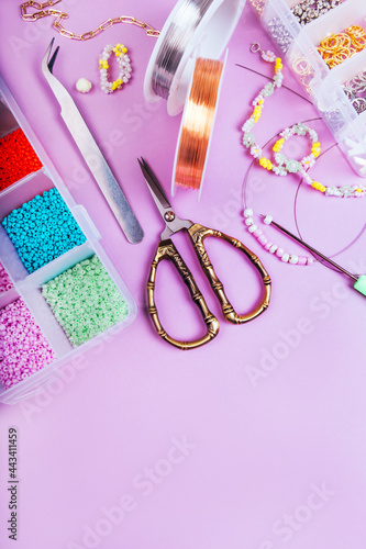 Creative flatlay of different seed and pearl beads with tools for making jewelry, wire string, scissors and necklace isolated on pink background.