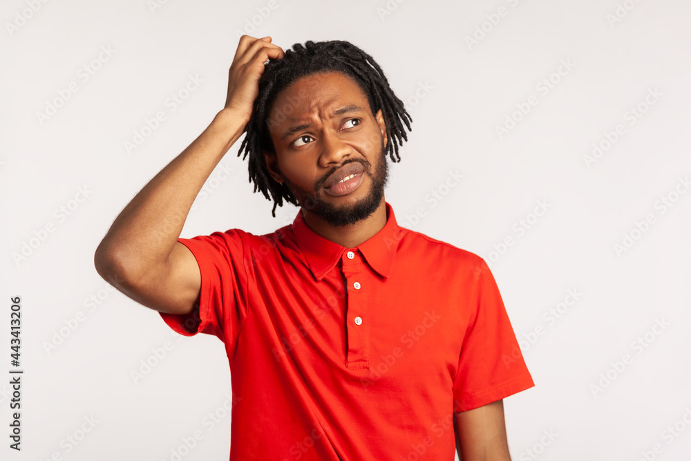 Doubt, need to think. Bearded man with dreadlocks wearing red casual style T-shirt, scratching head while thinking with confused puzzled face. Indoor studio shot isolated on gray background. Doubt, need to think. Bearded man with dreadlocks wearing red casual style T-shirt, scratching head while thinking with confused puzzled face. Indoor studio shot isolated on gray background.