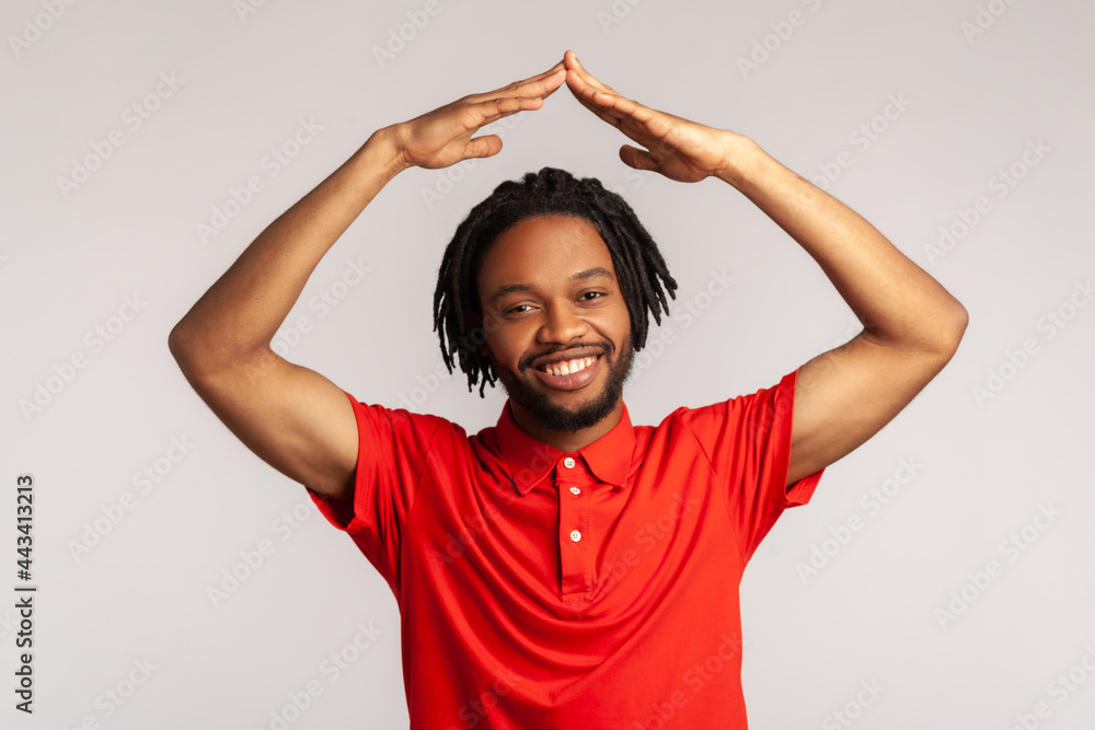 I'm in safety. Positive man with dreadlocks wearing red casual T-shirt, raising hands showing roof gesture and smiling contentedly, dreaming of house. Indoor studio shot isolated on gray background. I'm in safety. Positive man with dreadlocks wearing red casual T-shirt, raising hands showing roof gesture and smiling contentedly, dreaming of house. Indoor studio shot isolated on gray background.