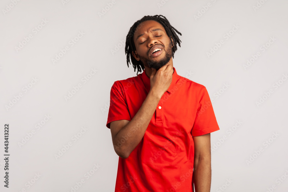 Portrait of bearded man with dreadlocks wearing red casual style T-shirt, touching painful neck, suffering sore throat, virus or tonsillitis. Indoor studio shot isolated on gray background. Portrait of bearded man with dreadlocks wearing red casual style T-shirt, touching painful neck, suffering sore throat, virus or tonsillitis. Indoor studio shot isolated on gray background.