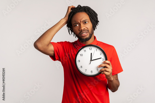 Confused man with dreadlocks wearing red T-shirt, rubbing his head holding in hands big wallclock, has no time , worried about deadline, looking away. Indoor studio shot isolated on gray background. photo