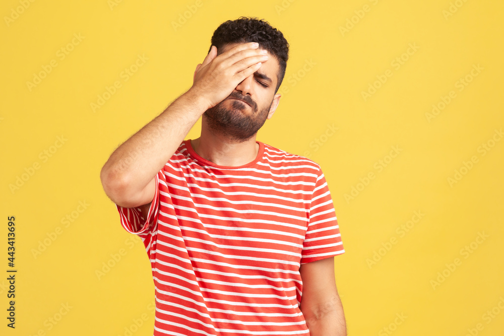 Unhappy forgetful man with beard in striped t-shirt making facepalm gesture keeping hand on head, blaming himself for bad memory, unforgivable mistake. Indoor studio shot isolated on yellow background Unhappy forgetful man with beard in striped t-shirt making facepalm gesture keeping hand on head, blaming himself for bad memory, unforgivable mistake. Indoor studio shot isolated on yellow background