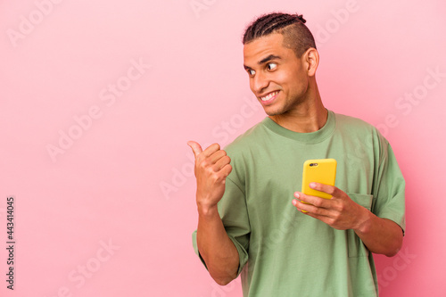 Young Venezuelan man holding a mobile phone isolated on pink background points with thumb finger away, laughing and carefree. © Asier