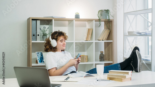 Lazy student. Homework break. Chilling time. Internet browsing. Happy sneaky overweight curly hair woman in headphones phone texting at modern light interior workplace. photo