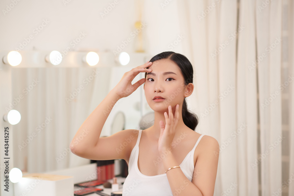 Beautiful young woman is touching her face and smiling while looking at the mirror Beautiful young woman is touching her face and smiling while looking at the mirror