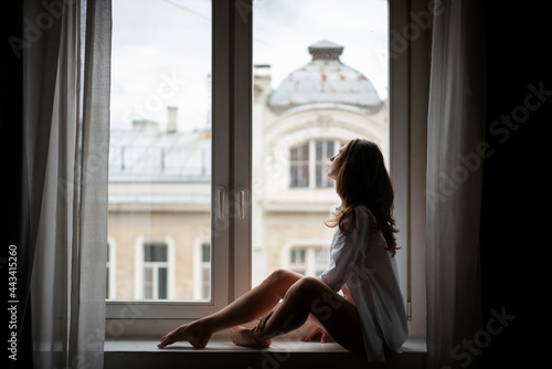 Beautiful sensual young woman with long hair in a white shirt elegantly sitting on the windowsill in an apartment with european city view