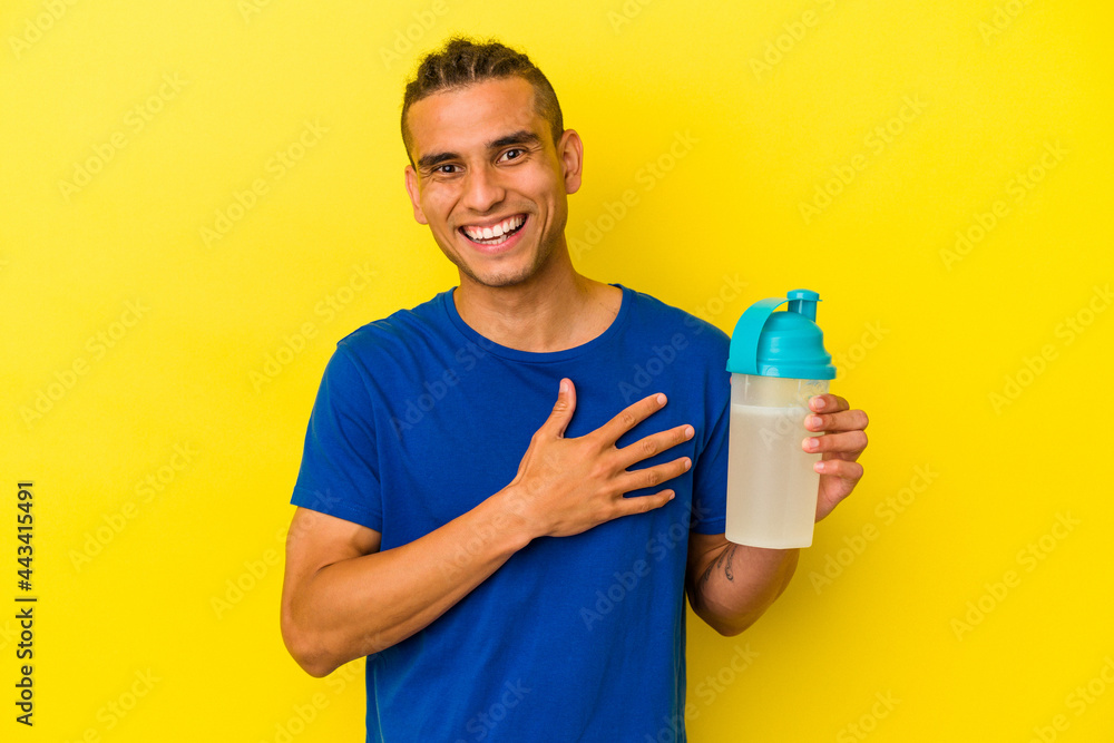 Young venezuelan man drinking a protein shake isolated on yellow background laughs out loudly keeping hand on chest. Young venezuelan man drinking a protein shake isolated on yellow background laughs out loudly keeping hand on chest.