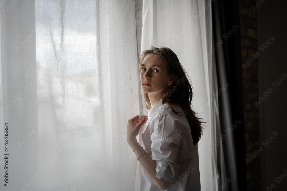 Beautiful sensual young woman with long hair in a white shirt standing by the window in the apartment