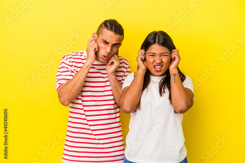 Young latin couple isolated on yellow background covering ears with hands.