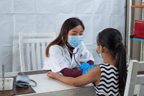 An asian doctor listening to the heartbeat of a child in her clinic. Doctor and patient little girl wearing facemasks. 