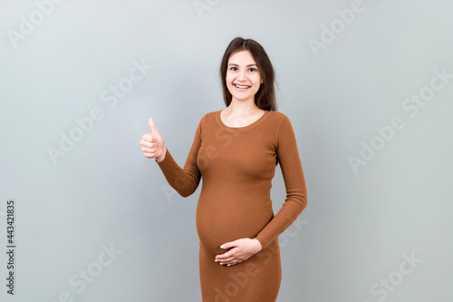 Happy pregnant woman showing thumb up cool gesture over her baby bump at Colored background. Happy maternity. Copy space