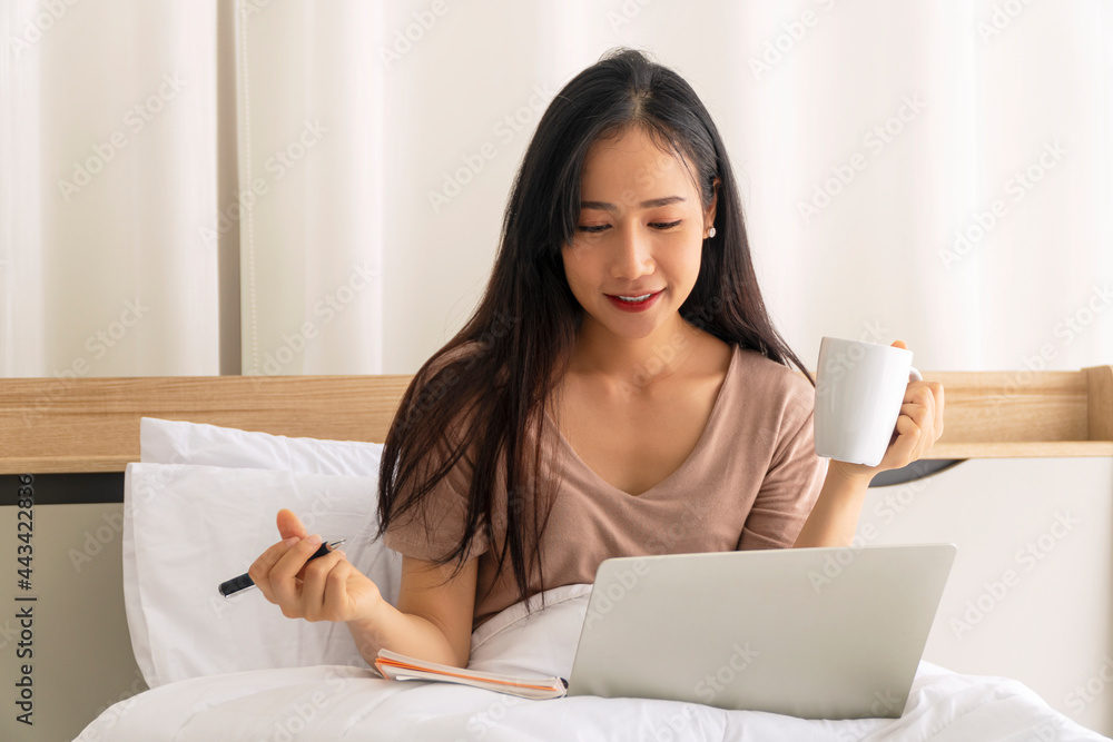 Freelance young woman using laptop working on internet holding cup coffee in own hand while sitting on white bed with pillow in the morning at apartment. Lifestyle work home with technology concept. Freelance young woman using laptop working on internet holding cup coffee in own hand while sitting on white bed with pillow in the morning at apartment. Lifestyle work home with technology concept.